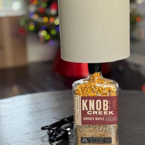 May include: A unique lamp made from a repurposed Knob Creek Smoked Maple bourbon bottle. The bottle is filled with grains and topped with a beige lampshade. The lamp features the brand's label and a black power cord.