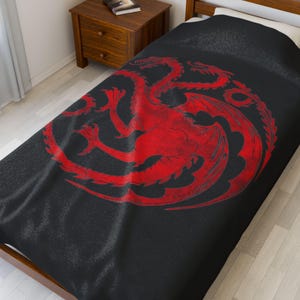 Könnte beinhalten: Schwarze Bettdecke mit einem großen, verwitterten roten Emblem eines dreiköpfigen Drachen. Die Bettwäsche ist über ein Bett drapiert. Ein Nachttisch aus Holz und ein Buch sind im Hintergrund zu sehen. Das Design ist auffällig.