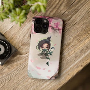 Könnte beinhalten: Eine Smartphone-Hülle mit einem Chibi-Ninja-Charakter mit dunklem Haar und grüner Maske vor einem Hintergrund aus fallenden rosa Kirschblüten und Zweigen, inklusive einer Aussparung für die dreifache Kameralinse an der Oberseite.