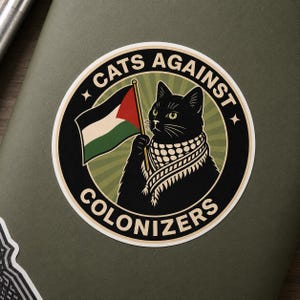 Puede incluir: Una pegatina circular con un gato negro que lleva un pañuelo keffiyeh y sostiene una bandera con franjas negras, blancas, verdes y rojas. El texto "CATS AGAINST COLONIZERS" está impreso en un borde circular sobre un fondo verde con rayos.