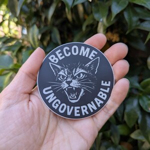 May include: A round, black sticker with white text and a cat illustration. The text reads "BECOME UNGOVERNABLE" around the image of a snarling cat. The sticker is held in a hand against a background of green foliage.