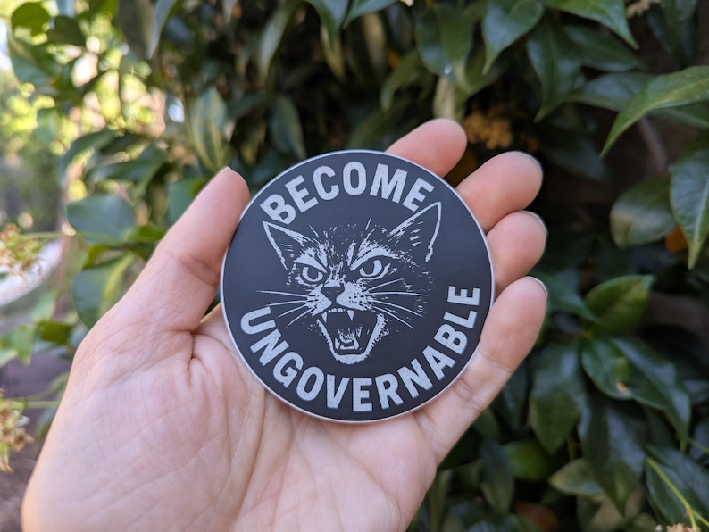 May include: A round, black sticker with white text and a cat illustration. The text reads "BECOME UNGOVERNABLE" around the image of a snarling cat. The sticker is held in a hand against a background of green foliage.