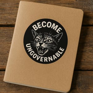 May include: A tan notebook with a black circular graphic featuring a snarling cat and the words "BECOME UNGOVERNABLE" in white. The notebook is on a wooden surface.
