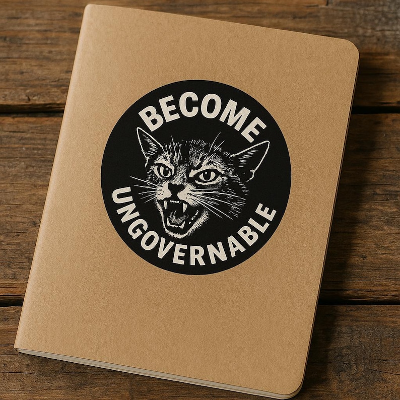 May include: A tan notebook with a black circular graphic featuring a snarling cat and the words "BECOME UNGOVERNABLE" in white. The notebook is on a wooden surface.