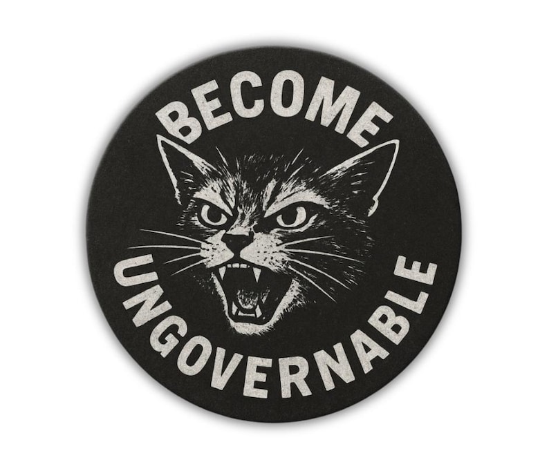 May include: Round black coaster with a white illustration of an angry cat's face and the words "BECOME UNGOVERNABLE" in a circular design. The cat is snarling with its mouth open, showing its teeth.