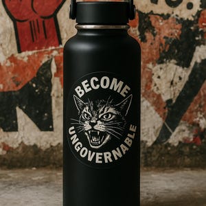 May include: Black water bottle with a black handle and a silver rim. The bottle has a white sticker with a snarling cat and the words "BECOME UNGOVERNABLE". The background is a distressed wall with a red fist.