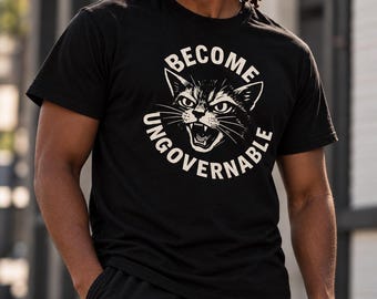 May include: A black short-sleeved t-shirt featuring a graphic of a snarling cat face in the center. The text "BECOME UNGOVERNABLE" is printed in a circular arrangement around the cat illustration in a light cream color on the black fabric.