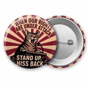 May include: A round pin-back button featuring a brown tabby cat at a podium with a microphone. The background has red and beige sunburst rays. Text reads: WHEN OUR RIGHTS ARE UNDER ATTACK STAND UP, HISS BACK. The back shows a metal pin mechanism.