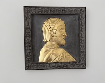 May include: A square wall plaque featuring a gold-colored relief of a bearded figure in profile, wearing a headpiece with a star emblem. The central figure is set against a dark background, framed by a border decorated with a repeating floral pattern.