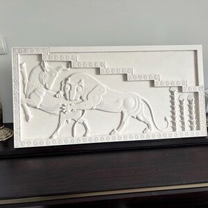 May include: White plaster relief sculpture depicting a lion attacking a bull. The sculpture is framed with a decorative border and set against a white background.