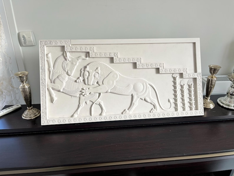 May include: White plaster relief sculpture depicting a lion attacking a bull. The sculpture is framed with a decorative border and set against a white background.