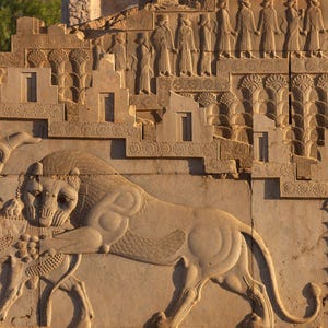 May include: A stone relief carving depicting a lion attacking a bull. The carving is on a wall with a background of stylized palm trees and a row of figures in relief.