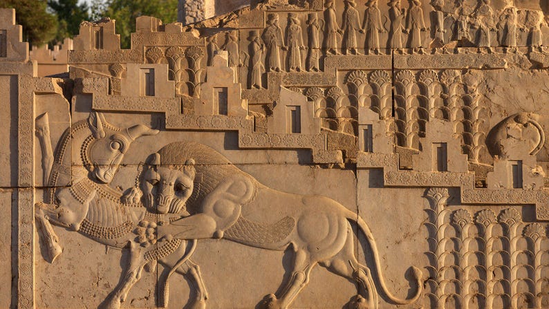 May include: A stone relief carving depicting a lion attacking a bull. The carving is on a wall with a background of stylized palm trees and a row of figures in relief.
