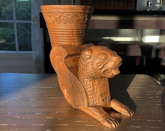 May include: A detailed wooden sculpture of a mythical creature, possibly a griffin, with a cup-like structure on its back. The sculpture is carved from light brown wood and features intricate details, including wings, a lion-like head, and clawed feet.