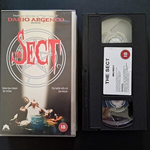 Può includere: Una videocassetta VHS e la sua custodia per il film horror "The Sect". La custodia presenta il titolo del film in rosso, un pentagramma e la scritta "Satana ha scelto le sue vittime". La videocassetta VHS ha un'etichetta con il titolo del film e una classificazione per età.