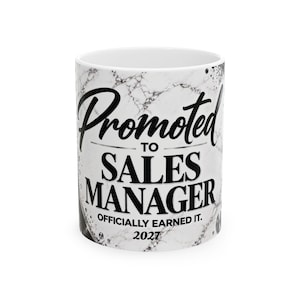 May include: A white ceramic mug with a black marble pattern featuring the black text Promoted TO SALES MANAGER OFFICIALLY EARNED IT. 2027. The mug has a cylindrical shape with a smooth finish and a clean white interior visible at the top rim.