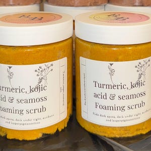 May include: Two jars of foaming scrub with white lids and labels. The labels read "Turmeric, kojic acid & seamoss Foaming scrub" with text below. The scrub is a bright yellow colour. The lids have a pink and orange design with the letters "B & B".