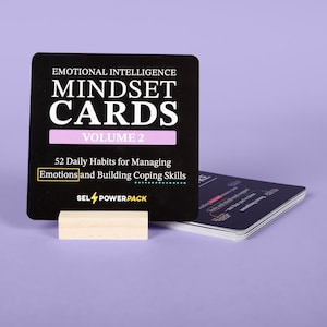 May include: A black card with white text that reads "EMOTIONAL INTELLIGENCE MINDSET CARDS VOLUME 2". The card is propped up by a small wooden block. The text "52 Daily Habits for Managing Emotions and Building Coping Skills" is also visible.