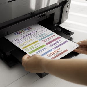 May include: A black printer printing a colorful page titled "GRATITUDE AT-A-GLANCE." The page lists ways to practice gratitude, including noticing the good, keeping a gratitude journal, and expressing thanks. The page is being held by a person.