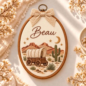 May include: An oval wooden embroidery hoop containing a fabric panel with the name Beau stitched in brown thread. The design features a desert landscape with a covered wagon, a cactus, a crescent moon, and stars, topped with a beige lace bow.