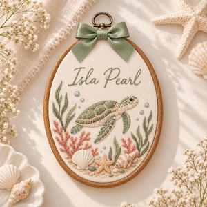 May include: An oval embroidery hoop featuring a sea turtle, coral, and seashells stitched on cream fabric. The name Isla Pearl is embroidered in green script above the turtle. A sage green satin ribbon bow is attached to the top metal hanger.
