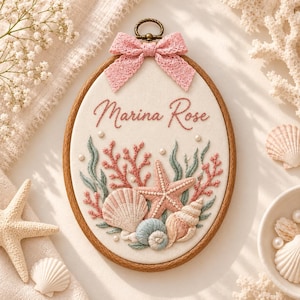 Puede incluir: Un bastidor de bordado ovalado con un panel de tela crema que tiene el nombre Marina Rose bordado en rosa. El diseño presenta coral rosa, algas verdes, una estrella de mar y conchas marinas, decorado con pequeñas perlas blancas y un lazo rosa.