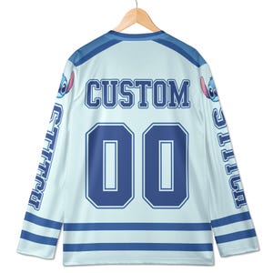 Puede incluir: Camiseta de hockey azul claro con la palabra "CUSTOM" y el n&uacute;mero "00" en azul marino. Las mangas tienen la palabra "STITCH" en azul marino. La camiseta presenta un personaje de dibujos animados en los hombros.