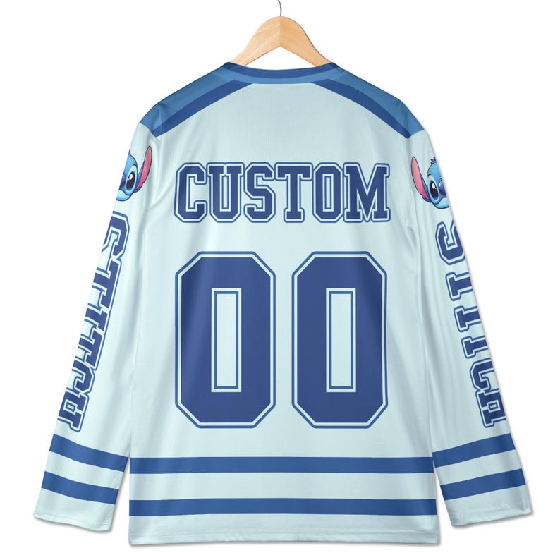 Puede incluir: Camiseta de hockey azul claro con la palabra "CUSTOM" y el n&uacute;mero "00" en azul marino. Las mangas tienen la palabra "STITCH" en azul marino. La camiseta presenta un personaje de dibujos animados en los hombros.