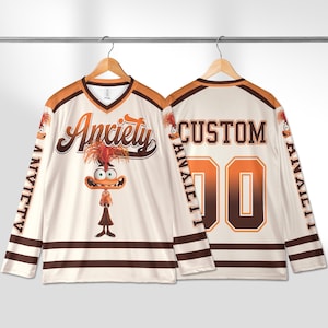 Puede incluir: Jersey de hockey crema con la palabra "Anxiety" en escritura marrón en la parte delantera, y un personaje de dibujos animados. La parte trasera presenta la palabra "CUSTOM" y el número "00" en marrón. Acentos marrones y naranjas.