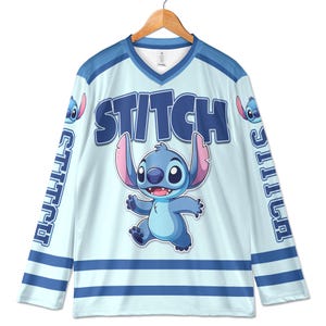 Puede incluir: Camiseta de hockey azul claro con la palabra "STITCH" en letras azules grandes y un personaje de dibujos animados. La camiseta tiene rayas azules y la palabra "STITCH" verticalmente en las mangas. La camiseta est&aacute; colgada en una percha de madera.