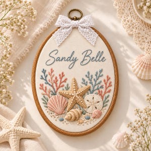 May include: An oval embroidery hoop containing a cream-colored fabric panel with the embroidered name Sandy Belle in blue script. The design features stitched coral, a central starfish, and various seashells in shades of pink and tan.