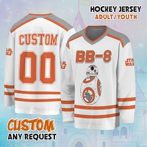 May include: A white hockey jersey featuring orange and grey accents, with the text BB-8 and a graphic of the BB-8 droid on the front. The back displays the word CUSTOM and the number 00 in orange. The sleeves feature the Star Wars logo.