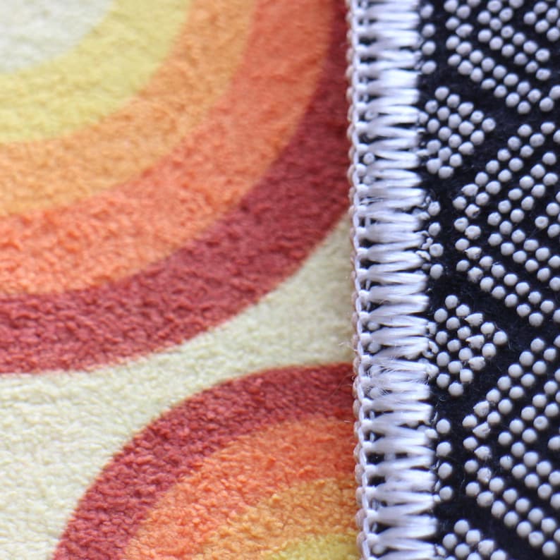 May include: Close-up of the edge of a rug with a colorful, abstract pattern. The rug has a white border and a black backing.