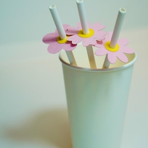 Könnte beinhalten: Ein weißer Pappbecher mit drei weißen Strohhalmen, die jeweils mit einer rosa Papierblume mit gelber Mitte versehen sind. Der Becher steht auf einer weißen Oberfläche.