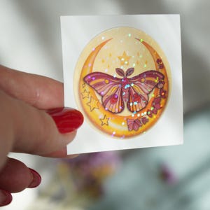 Könnte beinhalten: Ein runder Aufkleber mit einem Halbmond und einem rosa und orangefarbenen Schmetterling mit Glitzerakzenten. Der Aufkleber hat einen gelben Hintergrund mit kleinen weißen Sternen.