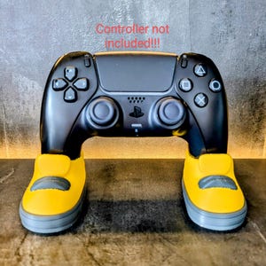 May include: A black PlayStation 5 controller stand with yellow and gray sneaker-shaped feet. The stand is designed to hold a controller. Text on the image reads "Controller not included!!!".