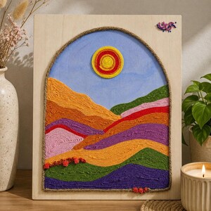May include: A colorful, handmade wall art piece featuring a landscape scene. The artwork depicts a mountain range with layers of orange, purple, green, and red yarn. A yellow and red sun is in a blue sky, framed by a jute rope arch.