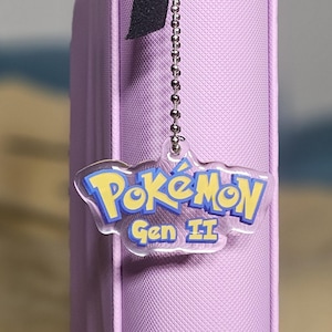 May include: A light purple binder with a black zipper and a black strap. A clear charm with the Pokemon logo and "Gen II" text hangs from a silver chain attached to the zipper.