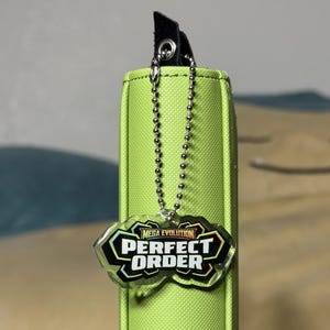 May include: A lime green binder with a black accent and a silver chain. The binder has a tag that reads "MEGA EVOLUTION PERFECT ORDER" in black and white. The binder is a storage item.