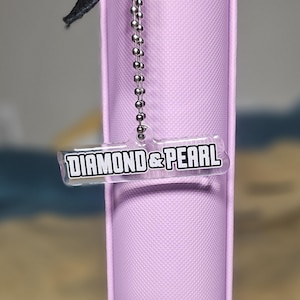 May include: A light purple, rectangular case with a black strap and a silver ball chain. A clear tag with the words "DIAMOND & PEARL" in a black, bold font hangs from the chain. The case has a smooth, matte finish.
