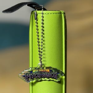 May include: A bright lime green binder with a textured surface features a silver metal ball chain attached to the spine. A clear acrylic charm hangs from the chain, displaying the text Mega Evolution Pitch Black in purple and yellow letters.