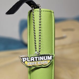 May include: A lime green card holder with a black strap and a silver chain. The holder has a clear tag with the words "PLATINUM ARCEUS" in white and yellow. The holder is likely for trading cards.