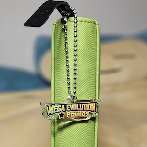 May include: A lime green rectangular case with a black fabric loop and a silver ball chain. A clear plastic charm with the words "MEGA EVOLUTION Promos" in a colorful design hangs from the chain.