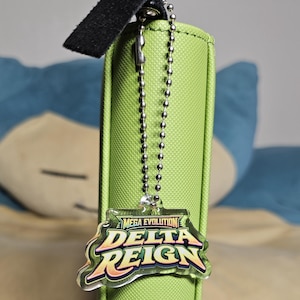 May include: A clear acrylic keychain featuring the text "MEGA EVOLUTION DELTA REIGN" in yellow, orange, and purple lettering. The keychain is attached to a silver-toned metal ball chain, which is secured to the spine of a lime green textured binder.