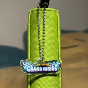 May include: A lime green cylindrical case with a black strap and a silver ball chain. A clear, rectangular charm with the words "MEGA EVOLUTION CHAOS RISING" in blue and white is attached to the chain.