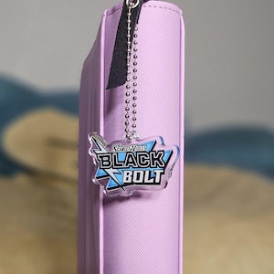 May include: A light purple, rectangular case with a black leather strap and a silver chain. A clear charm with the words "Scarlet & Violet BLACK BOLT" in white and blue hangs from the chain. The case has a zipper and a textured surface.