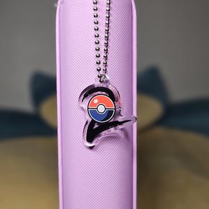 May include: A light purple pencil case with a black zipper pull and a silver ball chain. A clear acrylic charm with a red, white, and blue design hangs from the chain. The charm has a black outline.