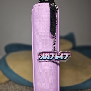 May include: A light purple card binder with a black zipper pull and a chain attached to a clear acrylic charm. The charm has Japanese text in pink, blue, and white. The binder is vertical and has a textured surface.