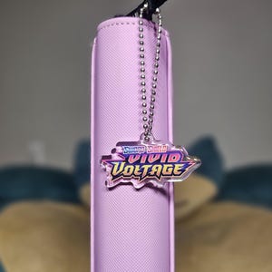 May include: A light purple card holder with a black strap and a silver chain. A clear charm with the words "Sword & Shield Vivid Voltage" in purple, pink, and yellow hangs from the chain.