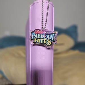 May include: A purple binder with a silver chain attached to a clear plastic charm with the text "Scarlet & Violet Paldean Fates" in colorful lettering.